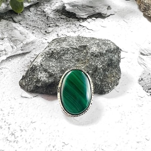 Könnte beinhalten: Ovale Malachit-Ring mit einer silberfarbenen, strukturierten Lünette. Der Ring hat einen leuchtend grünen Stein mit natürlichen Mustern. Der Ring ist vor einem strukturierten weiß-grauen Hintergrund mit einem großen grauen Felsen und grünem Laub platziert.