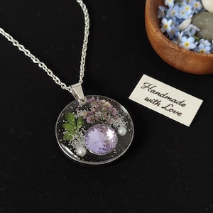 Könnte beinhalten: Halskette mit rundem Anhänger. Der Anhänger enthält getrocknete Blumen, einen lila Edelstein und Perlen in klarem Harz. Ein Etikett mit der Aufschrift "Handmade with Love".