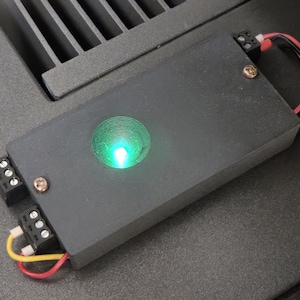 May include: A black electronic device with a bright green light. The device has connectors and wires attached. The rectangular device appears to be made of metal or a similar material. The device is on a dark surface.
