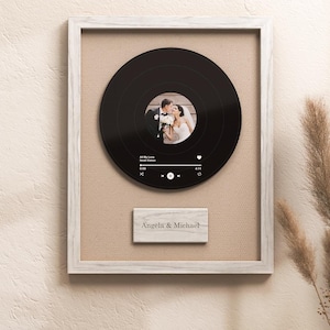 May include: A framed wedding gift featuring a black vinyl record with a photo of a couple. The record is set against a beige background within a white frame. A small wooden plaque below reads "Angela & Michael."