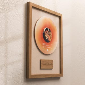 May include: A framed artwork with a vinyl record design, featuring a couple's photo. The record is orange and white, set within a wooden frame. A small wooden plaque below the record reads "Angela & Michael". The frame is made of wood.