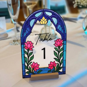 May include: A decorative table number with a stained-glass design. The arched frame is blue and purple, with a gold lotus flower at the top. Pink flowers and green leaves flank the number 1. The word "Table" is written in script.