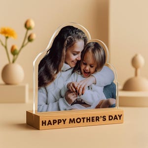 Könnte beinhalten: Ein transparenter Acryl-Bilderrahmen mit einem wolkenförmigen Ausschnitt, der ein Foto einer Mutter und ihrer Tochter zeigt. Der Rahmen steht auf einem Holzsockel mit dem eingravierten Text "HAPPY MOTHER'S DAY".