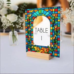 May include: A stained glass table number with a wooden base. The design features a colorful mosaic pattern with yellow flowers and green leaves. The text "TABLE 1" is displayed in a white space.