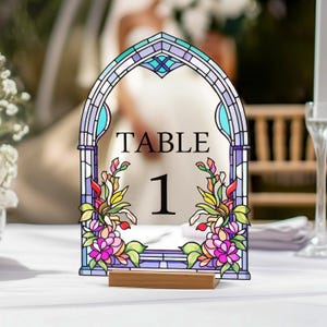 May include: A stained-glass style table number one. The arched design features a colorful floral arrangement and the word "TABLE" above the number 1. The base is a small, rectangular wooden block. The color palette includes purple, blue, green, and pink.