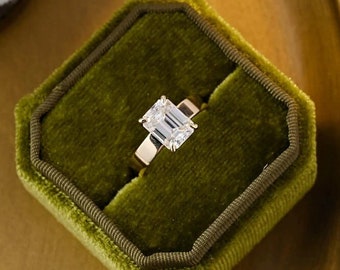 Puede incluir: Un anillo de oro con un gran diamante de talla esmeralda transparente engastado en una banda sencilla. El anillo se presenta en una caja de anillo de terciopelo verde.