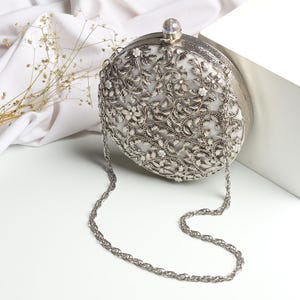 Puede incluir: Bolso de mano plateado, de forma ovalada, con intrincado diseño de filigrana floral y de hojas. El bolso tiene una delicada correa de cadena y un cierre con una parte superior de cristal. El fondo es blanco con una tela drapeada y ramitas de flores secas. Mide unos 20 cm.
