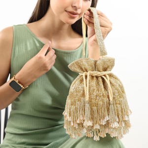 May include: A gold-colored drawstring handbag featuring an intricate beaded exterior with a shimmering metallic finish. The bag includes a single top handle, a thick braided cord closure, and a layered fringe base adorned with dangling bead accents.
