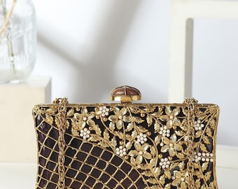 May include: An elaborate rectangular clutch bag with a gold chain strap. The bag has a dark purple interior, a gold filigree overlay, and is decorated with gold floral and white bead details. The clasp is a brown, oval-shaped stone.