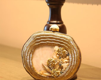 May include: A gold-coloured, round clutch bag with a decorative floral design. The clutch features a gold beaded border and a light pink centre. Gold roses and pearl-like beads adorn the front, creating an elegant accessory.
