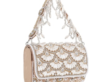 May include: A beige handbag featuring an intricate exterior adorned with white pearl-like beads, clear crystals, and gold-toned metallic accents arranged in a scalloped pattern. The handle is embellished with dangling pearl and crystal bead strands.