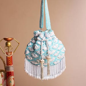 May include: A light blue beaded handbag with white floral accents and a white beaded fringe. The bag has a matching beaded handle and silver tassels. The bag is hanging from a silver hook.