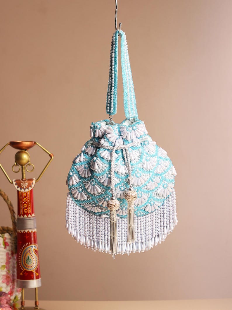 May include: A light blue beaded handbag with white floral accents and a white beaded fringe. The bag has a matching beaded handle and silver tassels. The bag is hanging from a silver hook.