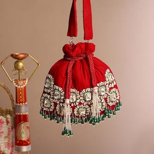 May include: A red velvet drawstring bag with a decorative design. The bag features a floral pattern with clear and green embellishments. It has a red handle and hanging tassels with green beads.