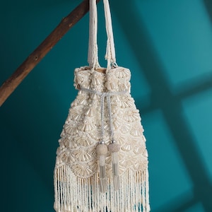 May include: Ivory beaded handbag with a drawstring closure and a long beaded handle. The bag is adorned with pearl-like beads and features a fringe along the bottom edge. Two decorative tassels hang from the drawstring. The bag is hanging from a tree branch.