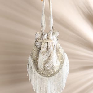 Puede incluir: Un bolso con cordón y cuentas de color blanco roto con una larga borla. El bolso presenta un patrón detallado de cuentas plateadas y blancas, con detalles de perlas. Tiene dos correas blancas y está colgado de una percha de madera.