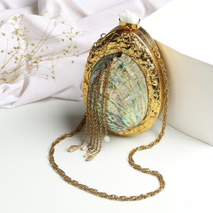 May include: A gold-colored, oval-shaped clutch with a textured surface and a chain strap. The clutch features an iridescent shell design in the center and is adorned with dangling gold chains and decorative beads. A white stone sits atop the clutch.