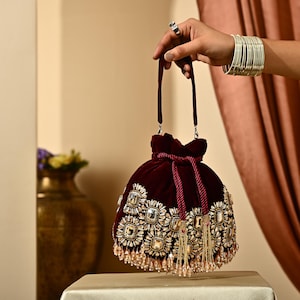 May include: A burgundy velvet drawstring bag with ornate gold and crystal embellishments. The bag features a tassel fringe and a matching burgundy drawstring closure. The bag is held by a hand, showcasing its elegant design.