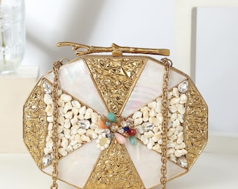 May include: A decorative gold clutch bag with a chain strap. The bag has a geometric design with white and gold sections, adorned with small white stones and colourful beads. A branch-shaped clasp is at the top.