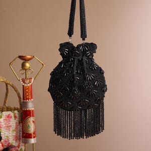 May include: A black beaded handbag with a drawstring closure and beaded fringe. The bag has two beaded handles and is suspended from a silver hook. The bag's surface is covered in intricate beadwork, creating a textured appearance.