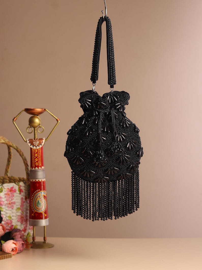 May include: A black beaded handbag with a drawstring closure and beaded fringe. The bag has two beaded handles and is suspended from a silver hook. The bag's surface is covered in intricate beadwork, creating a textured appearance.