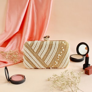 Op de afbeelding: Een rechthoekige clutch met een gouden frame en een parel- en goudkralenontwerp. De clutch heeft een decoratieve sluiting en is omgeven door make-up items, waaronder blush, poeder en lippenstift. De achtergrond heeft een roze stof.