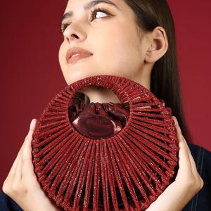 Op de afbeelding: Een rode, ronde handtas met een gestructureerd, geribbeld ontwerp. De tas heeft een handvat aan de bovenkant en een glanzende, metallic binnenkant. De tas wordt door een persoon tegen een rode achtergrond omhoog gehouden.