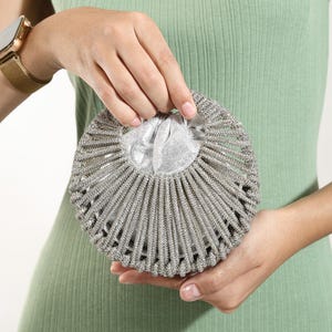 Op de afbeelding: Een ronde zilveren handtas met een stijf, waaiervormig buitenframe bedekt met kleine, sprankelende kristallen. De tas heeft een zachte, metallic zilveren stoffen binnenzak die aan de bovenkant samentrekt, wat een contrast vormt met de gestructureerde, stralende, met kristallen ingelegde buitenkant.