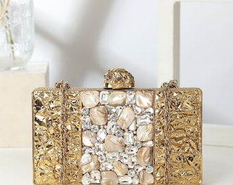 May include: A gold-coloured clutch bag with a textured surface and a chain strap. The centre is decorated with a mosaic of iridescent stones and clear crystals. The clasp is a gold-toned, textured sphere.