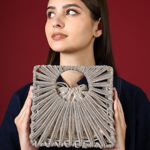 Puede incluir: Un bolso de mano cuadrado, de color dorado, con una abertura circular y un cierre de cordón. El bolso está hecho de un material tejido con un diseño de rayos de sol. El fondo es de color granate.