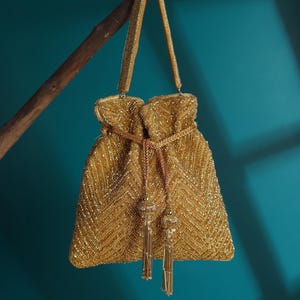 May include: A gold beaded drawstring bag with a chevron pattern. The bag has a matching beaded handle and decorative tassels. The bag is hanging from a brown branch against a teal background.