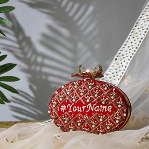 Könnte beinhalten: Eine rote, ovale Clutch mit aufwendiger goldener Stickerei und Perlenakzenten. Auf der Clutch steht der Text "#YourName" in weißer Schrift. Ein goldfarbener Verschluss mit einer Perle befindet sich oben. Die Clutch liegt auf cremefarbenem Stoff.