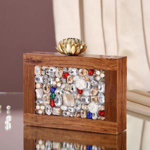 Può includere: Una scatola rettangolare in legno con un pannello frontale decorativo. Il pannello è adornato da una serie di abbellimenti chiari, colorati e simili a perle. Un accento floreale color oro si trova sopra la scatola.