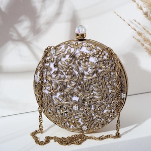 Peut inclure: Une pochette ronde ornée d'un cadre et d'une chaîne dorés. La pochette présente des motifs floraux et cachemire dorés complexes, avec un intérieur blanc. Un bijou clair et facetté orne le fermoir.