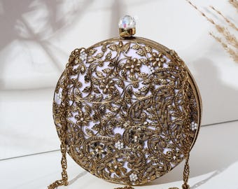 May include: A round, ornate clutch with a gold-colored metal frame and chain strap. The clutch features intricate floral and paisley designs in gold, with a white interior. A clear, faceted gem tops the clasp.