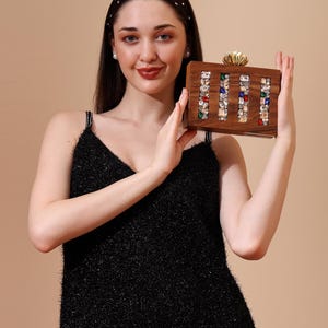 Op de afbeelding: Een rechthoekige houten clutch met kleurrijke edelstenen gerangschikt in verticale kolommen. De clutch heeft een goudkleurige sluiting en wordt vastgehouden door een persoon die een zwarte jurk draagt.