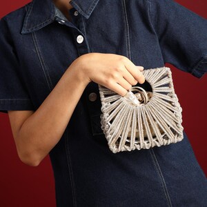 May include: A structured handbag featuring a metallic silver, cage-like exterior with a radial, fan-shaped pattern of corded strands. The bag has a circular cutout handle and contains a beige fabric drawstring pouch insert visible through the open frame.
