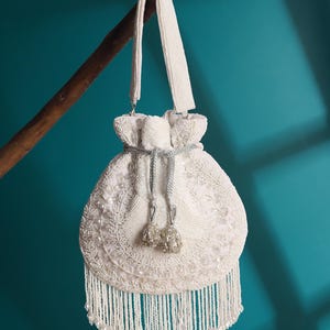 May include: A white beaded drawstring bag with a matching handle. The bag features intricate beadwork and a fringe of white beads along the bottom. Silver-colored tassels hang from the drawstring closure. The bag is suspended against a teal background.