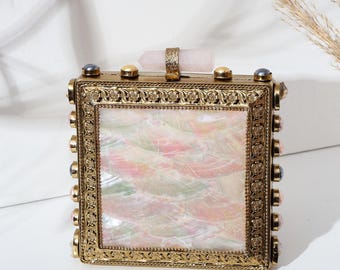 May include: A rectangular, gold-toned clutch with a mother-of-pearl front panel. The frame is adorned with decorative detailing and gemstone accents. A rose quartz crystal handle adds a unique touch to this elegant accessory.