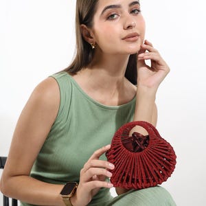 May include: A red, fan-shaped handbag with a structured design. The bag is held by a person wearing a green dress and a gold watch. The bag's design features a series of vertical lines radiating from the centre.