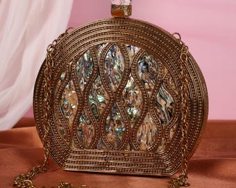May include: A gold-toned, circular clutch bag with an iridescent, patterned centre. It features a chain strap and a crystal-topped clasp. The design includes detailed metalwork and a decorative, swirling pattern.