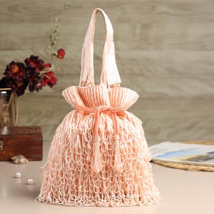 Puede incluir: Un bolso de mano color melocotón con cuentas, cierre de cordón y fondo de flecos. El bolso tiene un asa hecha del mismo material que el cuerpo. El bolso está sobre una mesa.