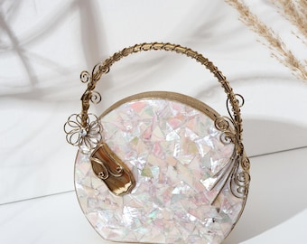 May include: A round, iridescent clutch bag with a gold handle and decorative features. The bag has a mosaic pattern of white and pastel pieces. A gold flower and clasp are on the front.