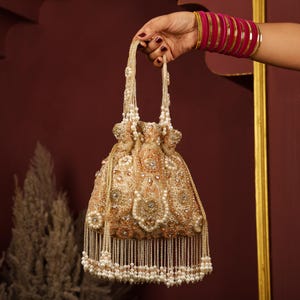 Puede incluir: Un bolso dorado con cordón, adornado con intrincados abalorios y detalles de perlas. El bolso presenta una borla de cuentas y delicadas asas. El diseño incluye patrones florales y adornos brillantes, creando una estética lujosa.