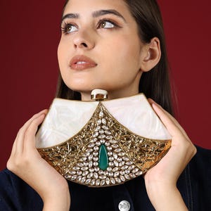 May include: A white and gold clutch with a decorative design. The clutch features a gold frame with clear crystals and a large green gemstone. The top of the clutch is white with a pearlescent finish.