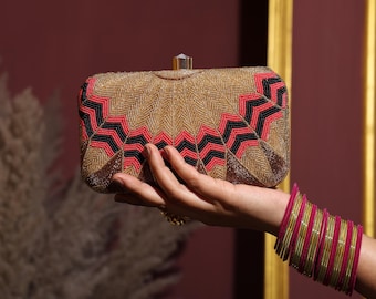 May include: A rectangular gold clutch with a geometric pattern in red and black. The clutch is held in a hand, with a stack of pink and gold bangles on the wrist. The clutch has a crystal clasp.