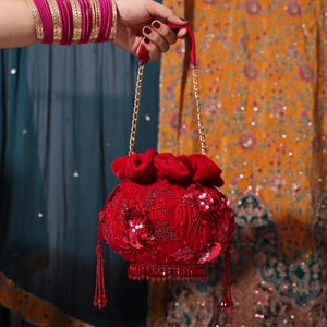 Może przedstawiać: Czerwona aksamitna torebka ze sznurkiem, złotymi łańcuszkami i koralikowymi frędzlami. Torebka jest ozdobiona cekinami i kwiatowymi aplikacjami. Ręka trzymająca torebkę jest ozdobiona różowymi bransoletkami.