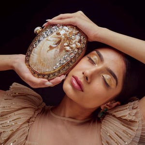 May include: An oval-shaped, gold-toned clutch with a pearl and crystal embellishment. The clutch features a shell-like design with floral accents. The frame is detailed with intricate patterns. The background is black.