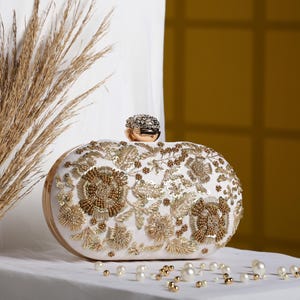 Peut inclure: Pochette ovale ivoire ornée de broderies et de perles dorées. Elle est dotée d'un fermoir orné de bijoux et d'une monture dorée. Des perles éparpillées ajoutent une touche d'élégance. Le design est floral et orné.