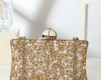 May include: A gold-coloured rectangular clutch bag with a floral pattern. The bag has a white interior and is decorated with gold leaves, flowers, and small white beads. A gold chain strap is attached. The clasp is white and gold.